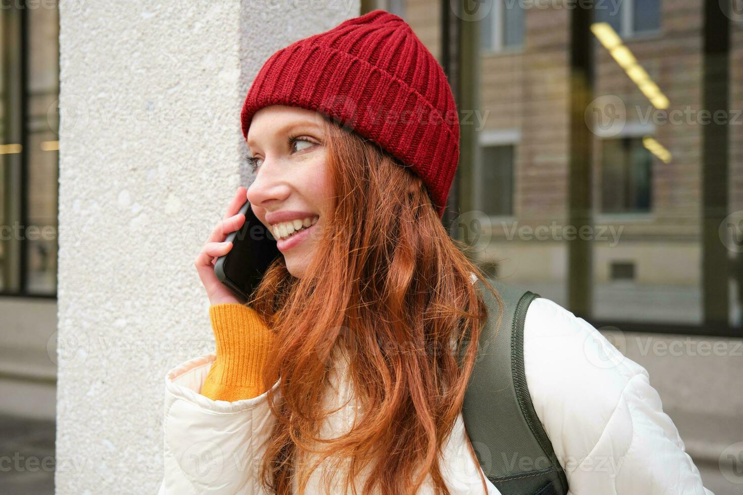 feliz lindo garota, ruiva turista fala em Móvel telefone, fazer uma ligar fora do país, tem conversação em Smartphone, em pé com mochila ao ar livre foto