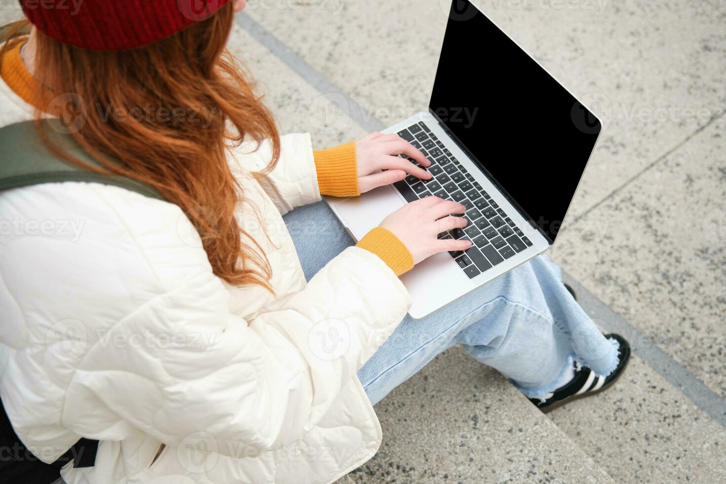 traseiro Visão do menina aluna digitando em computador portátil teclado, em branco Preto tela para local na rede Internet inscrição anúncio, sentado em escadas ao ar livre, conecta para público Wi-fi foto