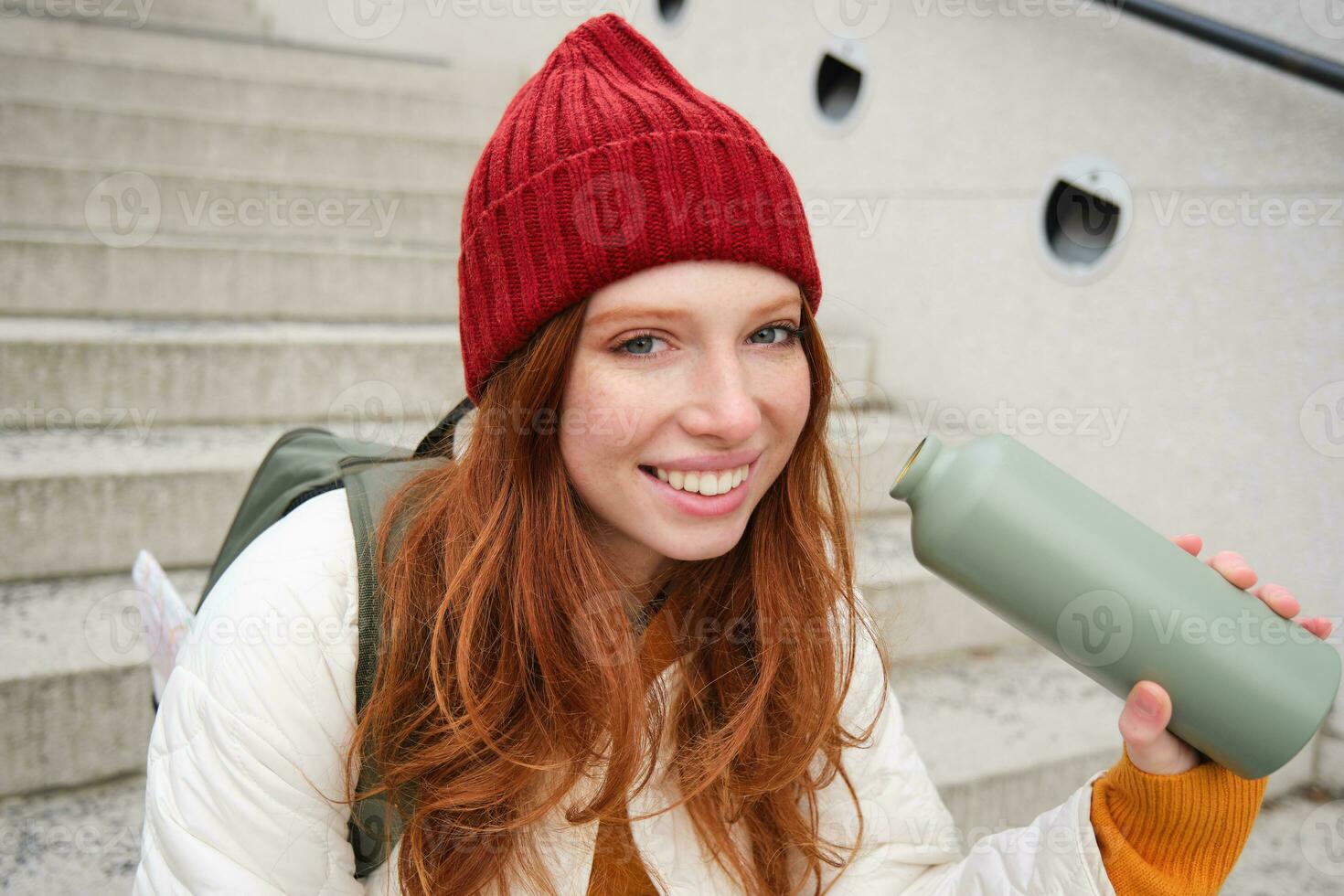 feliz ruiva mulher, turista com garrafa térmica, bebidas dela quente chá, café a partir de viagem frasco, testes de descanso durante dela viajando dentro cidade e passeios turísticos foto