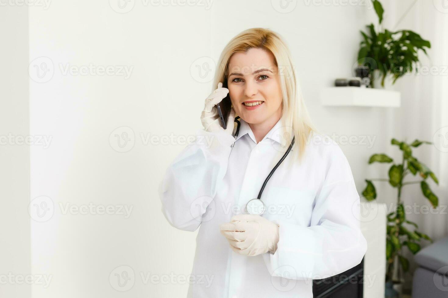 lado perfil retrato sorridente fêmea doutor, cuidados de saúde profissional dentro branco laboratório casaco com estetoscópio, analisando dados resultados em Móvel inteligente telefone em pé dentro hospital corredor corredor foto