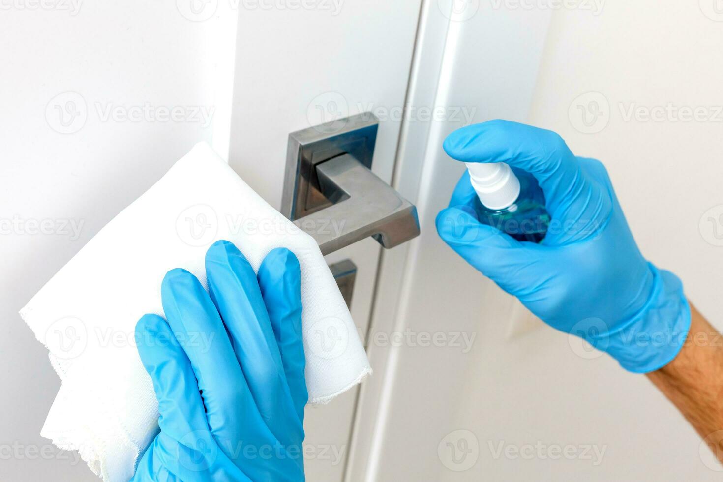 mãos dentro protetora luvas - 1 detém a pulverizador com a antisséptico, a de outros - lenços a porta lidar com com uma pano umedecido com uma desinfetante solução foto