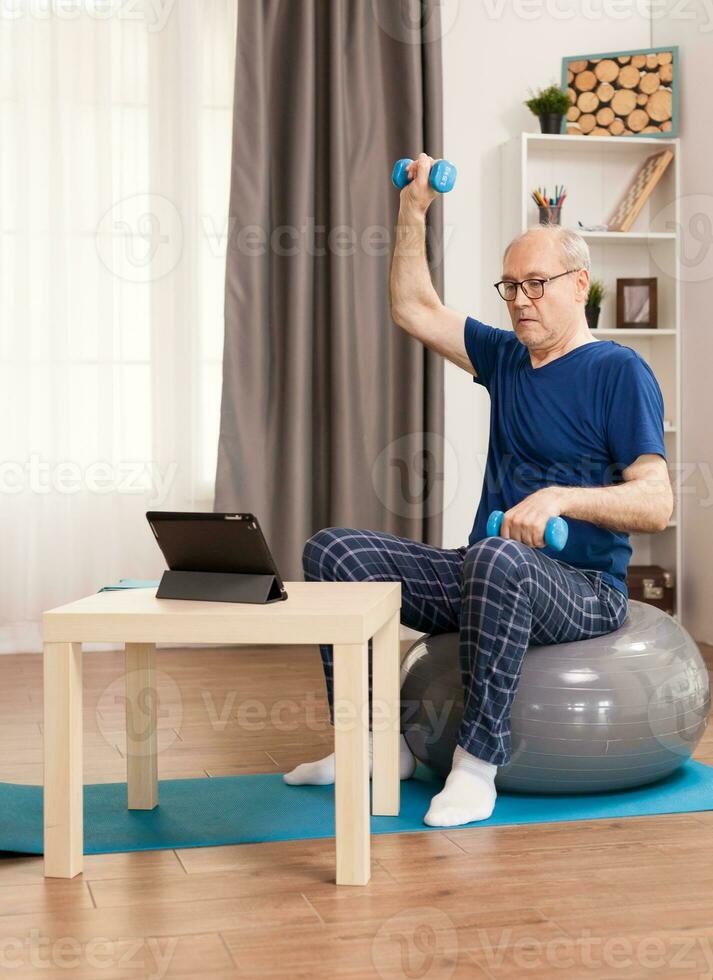 Senior homem fazendo músculo recuperação exercícios sentado em uma suíço bola dentro frente do a tábua. velho pessoa pensionista conectados Internet exercício Treinamento às casa esporte atividade com haltere, resistência banda, suíço bola às idosos aposentadoria idade. foto