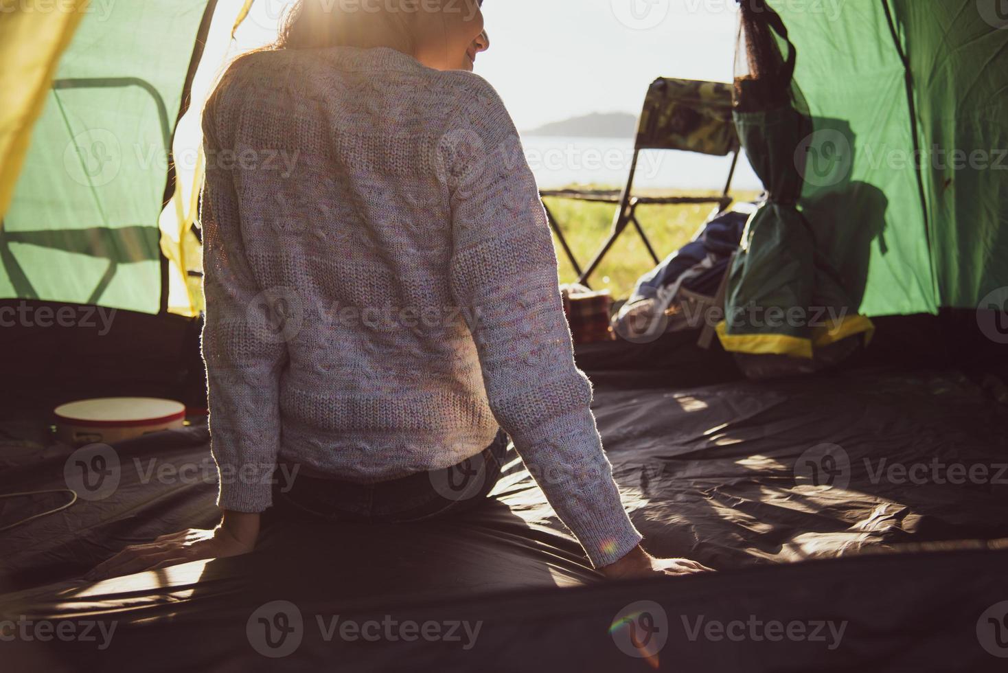 vista traseira do turista feminino feliz relaxando na barraca de acampamento com a montanha e o sol flare fundo. conceito de pessoas e estilos de vida. viagens e férias no prado ao ar livre. tema turismo e caminhadas foto