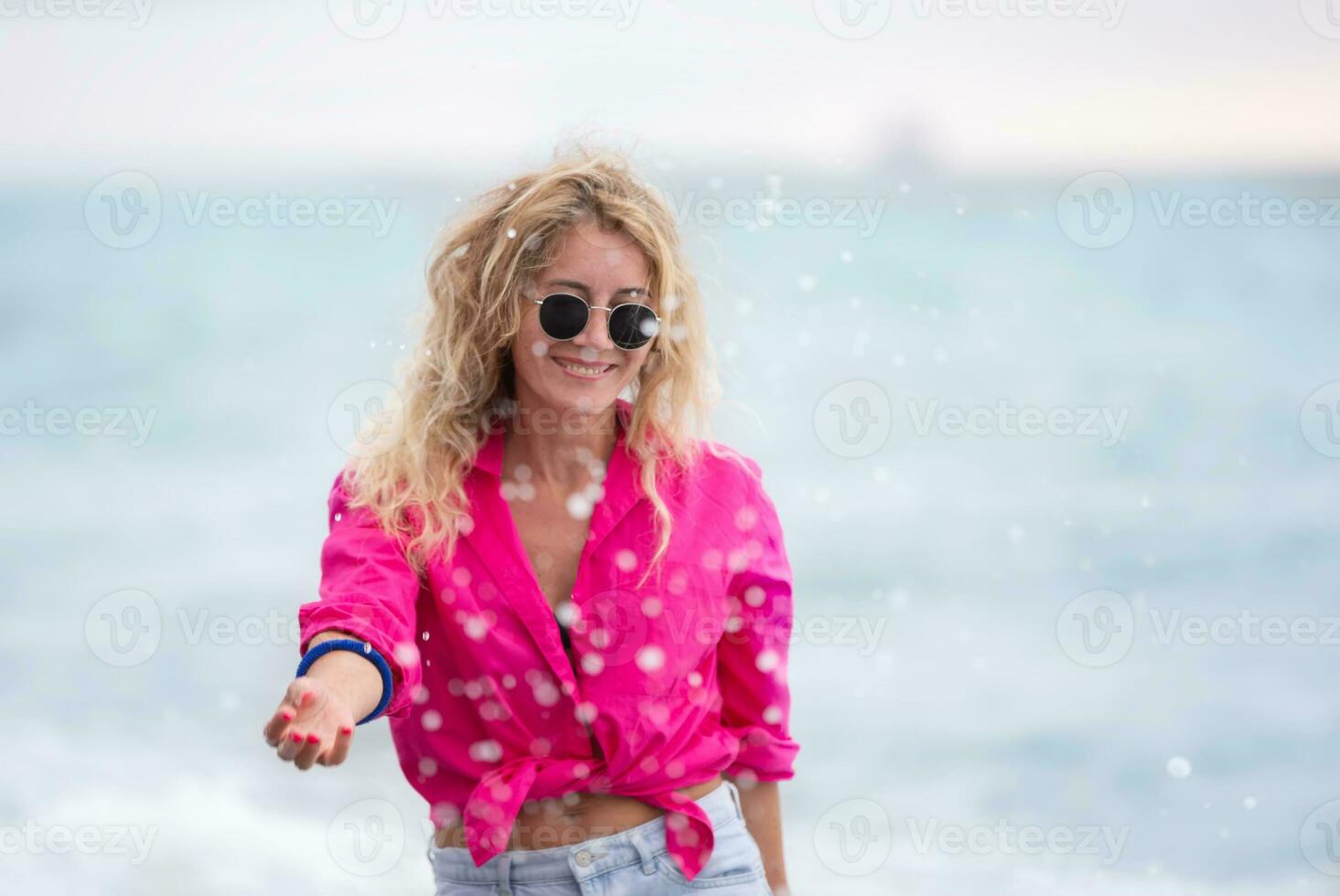 uma lindo mulher dentro oculos de sol e uma Rosa camisa salpicos mar água. foto