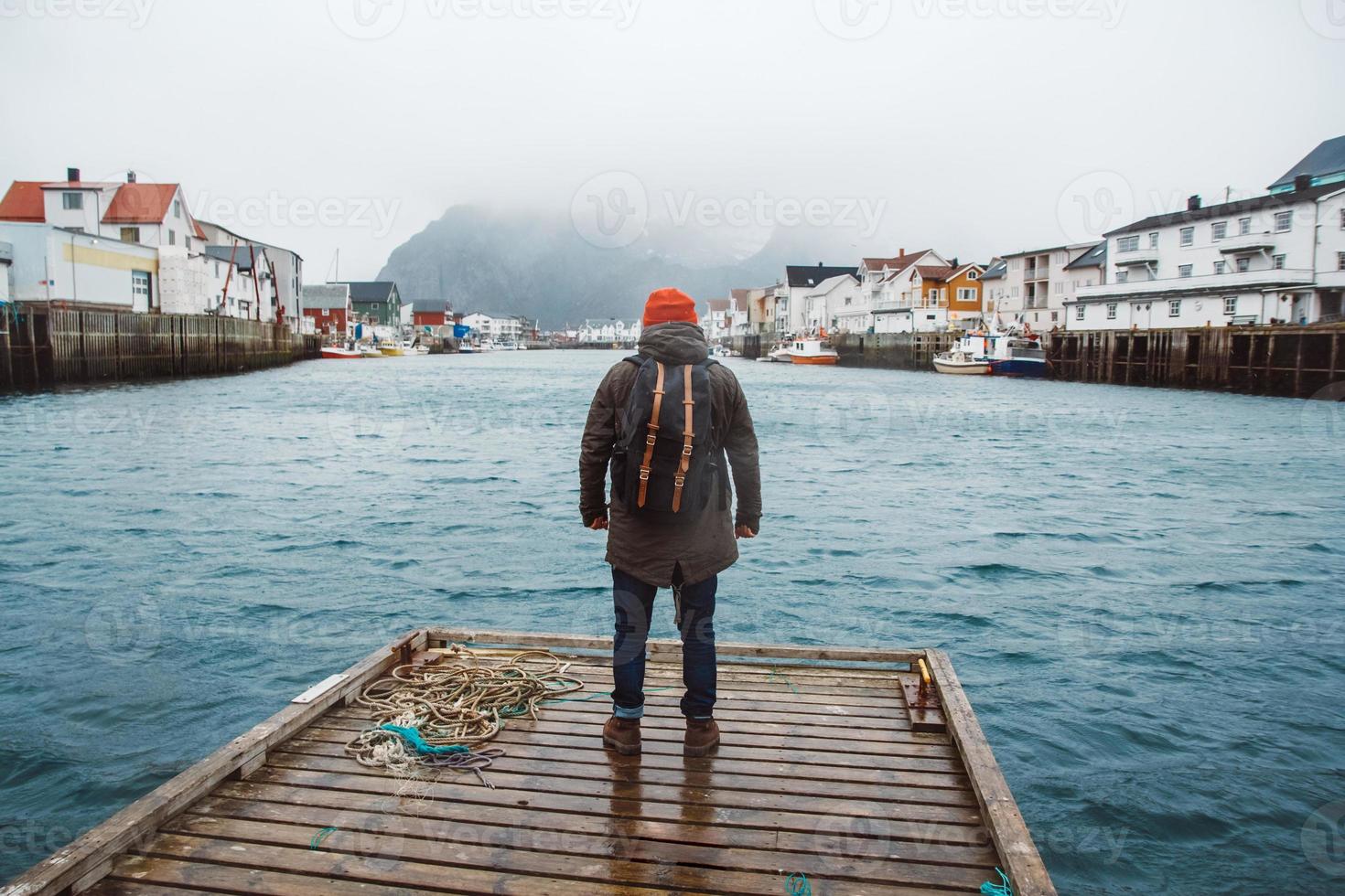 homem viajante com uma mochila em pé no cais de Woode no fundo de casas de pesca, montanha e lago foto