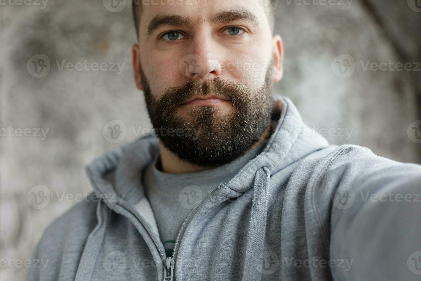 barbudo homem em uma cinzento fundo faz uma selfi foto