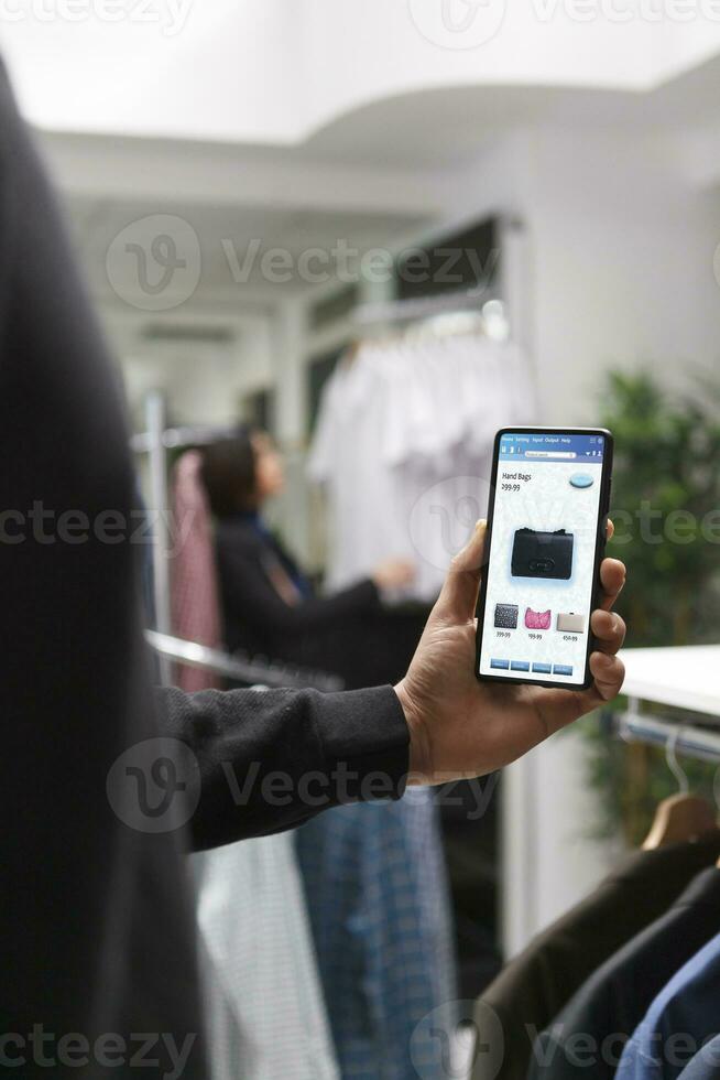 seletivo foco do caucasiano cliente segurando digital dispositivo dentro moderno boutique. à moda masculino comprador navegando para elegante mercadoria dentro roupas loja com assistência a partir de moderno tecnologia. foto