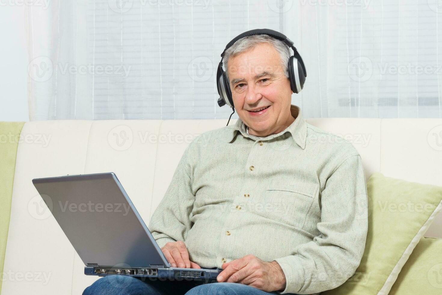 Senior homem sentado em sofá com fones de ouvido e usando computador portátil. foto