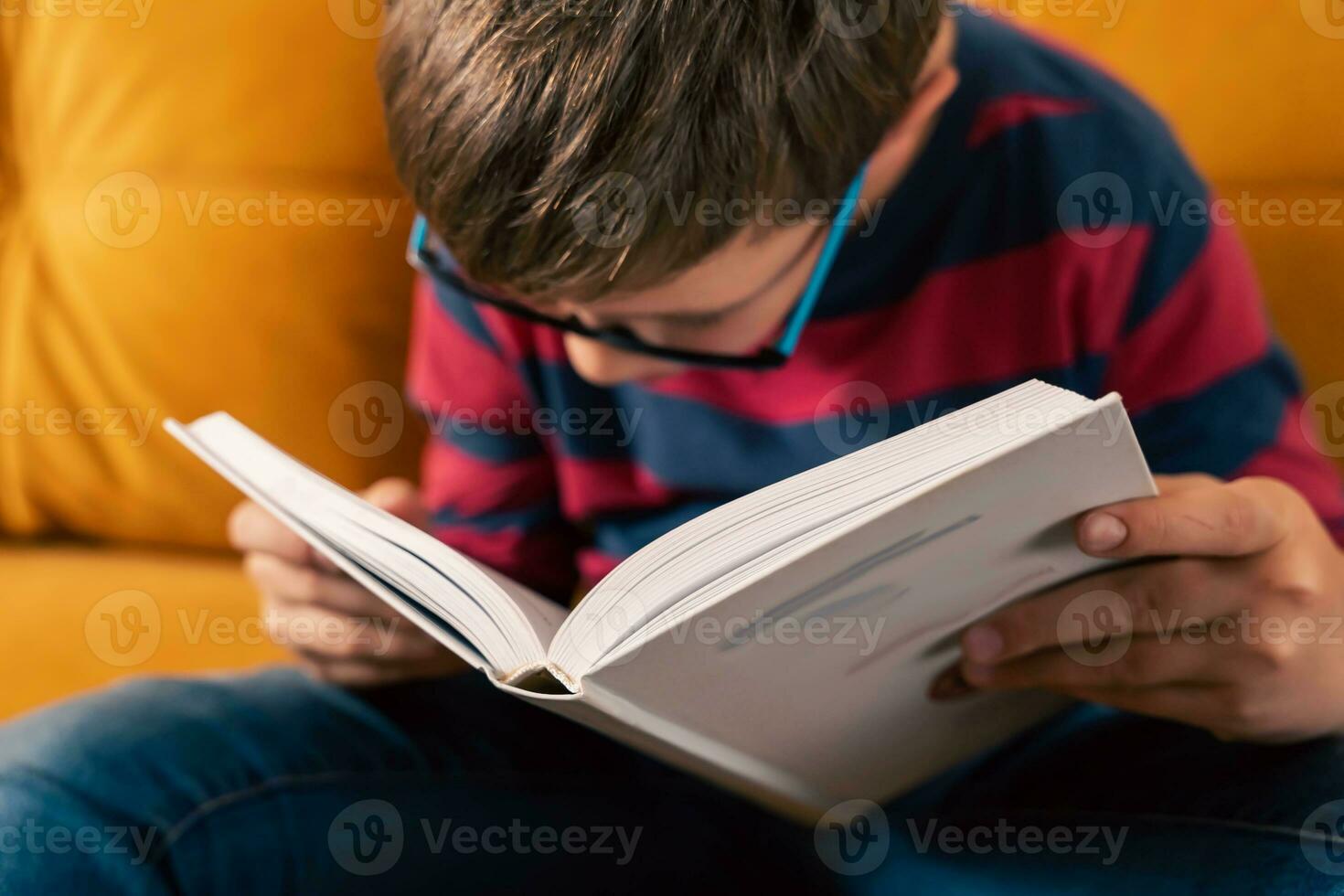 inquisitivo idade escolar Garoto com óculos lendo uma livro em a sofá foto