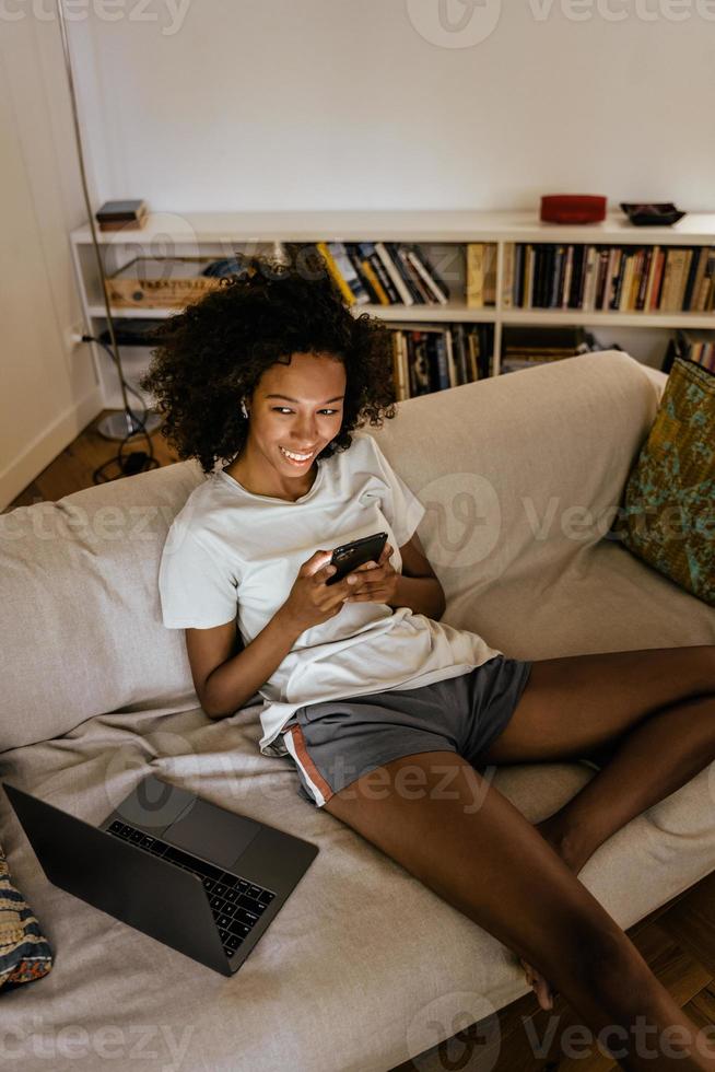 jovem negra com fones de ouvido usando o celular enquanto está descansando no sofá foto