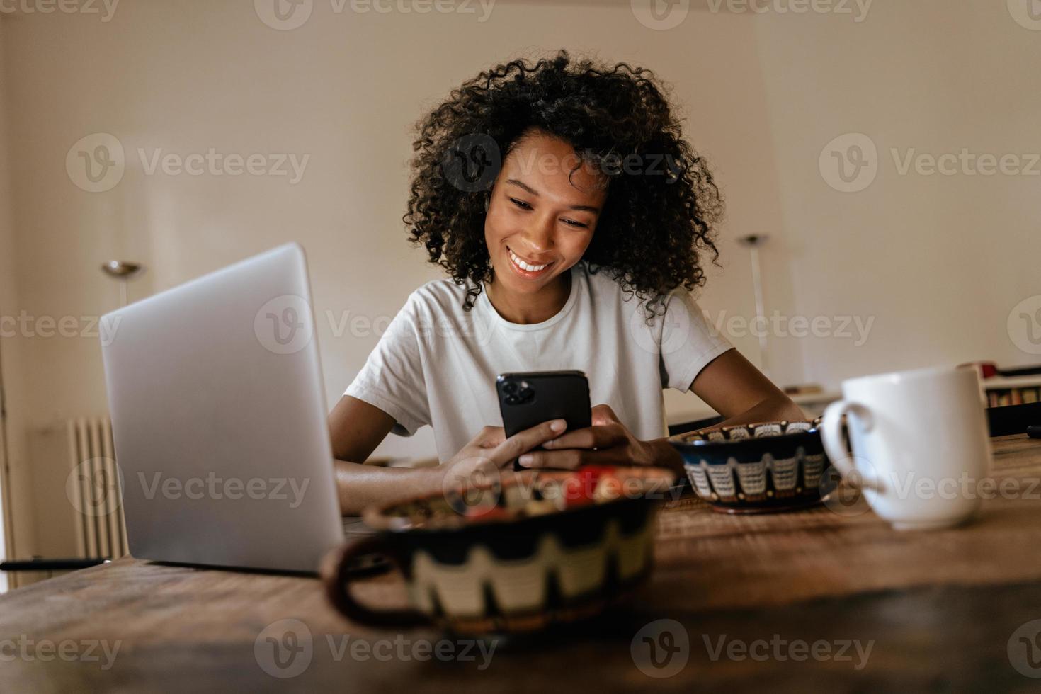 jovem negra usando telefone celular e laptop enquanto toma o café da manhã foto
