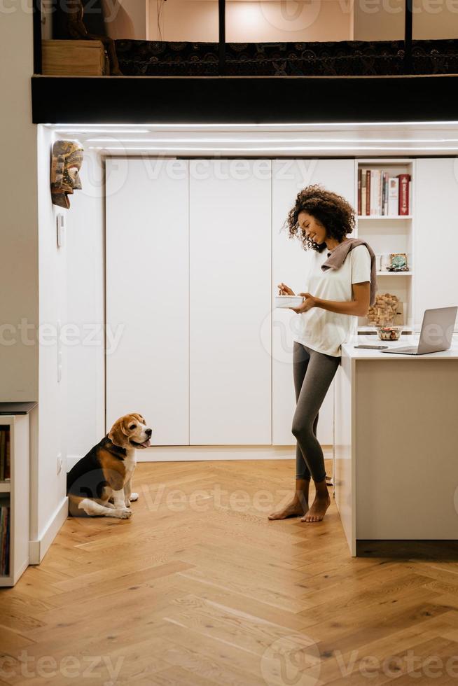 jovem negra tomando café da manhã enquanto olha para o cachorro na cozinha foto