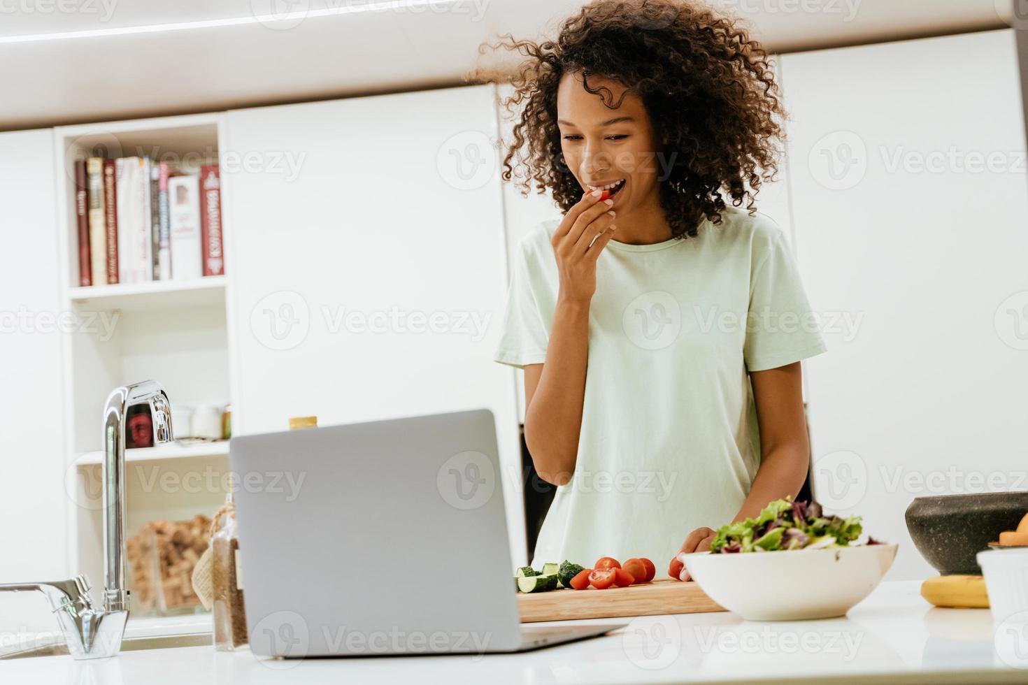 jovem negra fazendo salada enquanto usa o laptop na cozinha foto