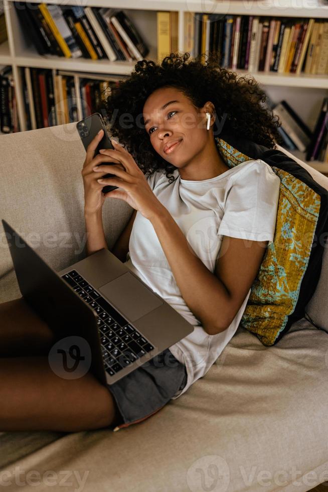 jovem negra usando telefone celular e laptop enquanto descansa no sofá foto