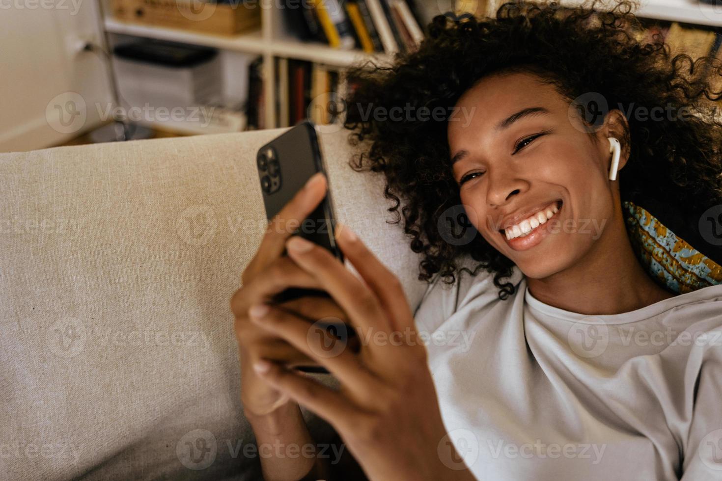 jovem negra com fones de ouvido usando o celular enquanto está descansando no sofá foto