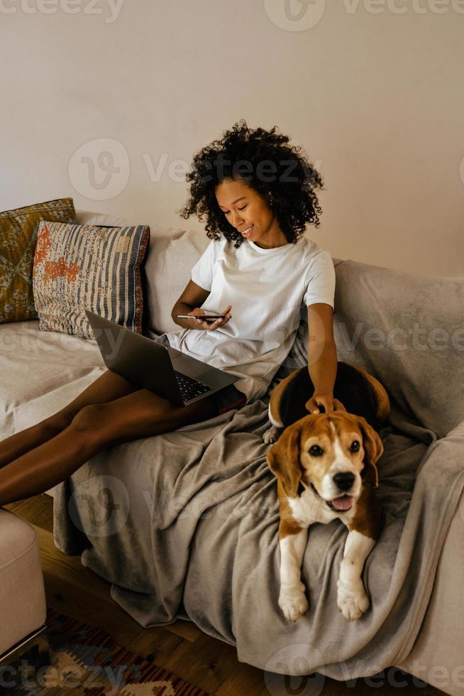 jovem negra usando o celular e acariciando o cachorro no sofá foto
