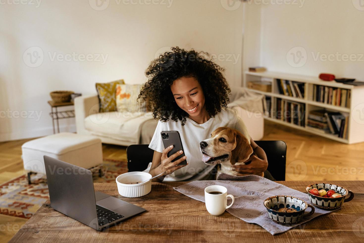 mulher negra usando celular e abraçando o cachorro enquanto tomava o café da manhã foto