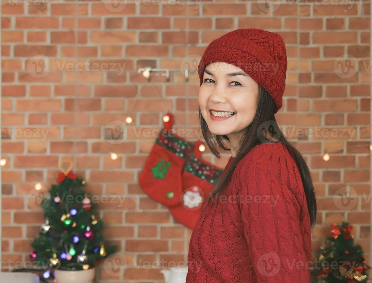 ásia mulher vestindo vermelho tricotado suéter e chapéu em pé dentro a cozinha com Natal decorações, sorridente alegremente e olhando às Câmera. inverno e Natal. foto