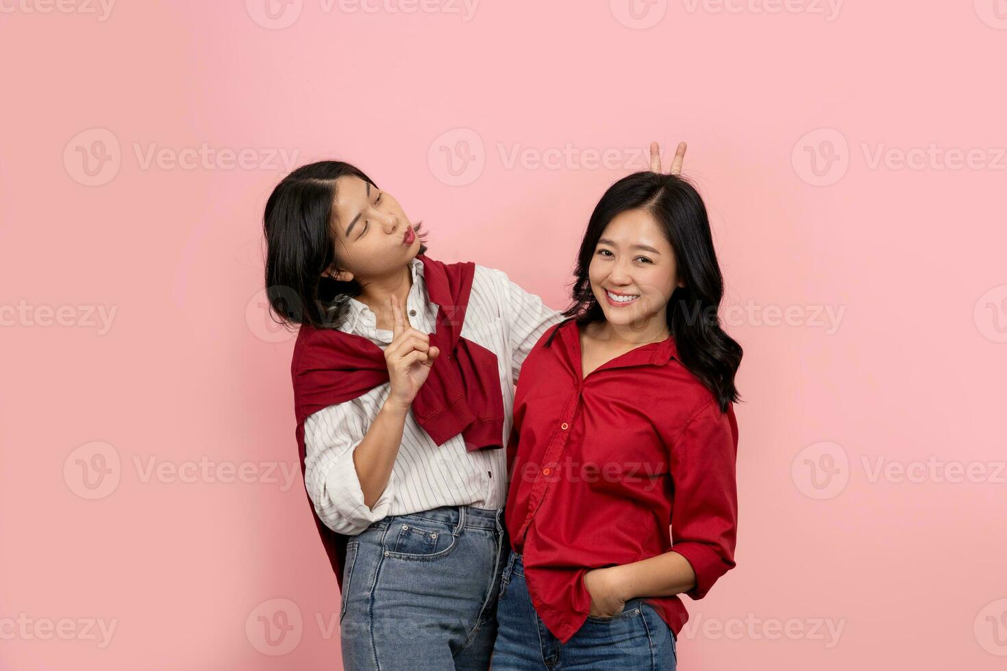 ásia fêmea amigos rindo e posando em Rosa fundo uma lindo sorridente mulher vestindo uma vermelho camisa é feliz. uma alegre menina vestindo uma branco camisa dá Coelho orelhas para amigo foto