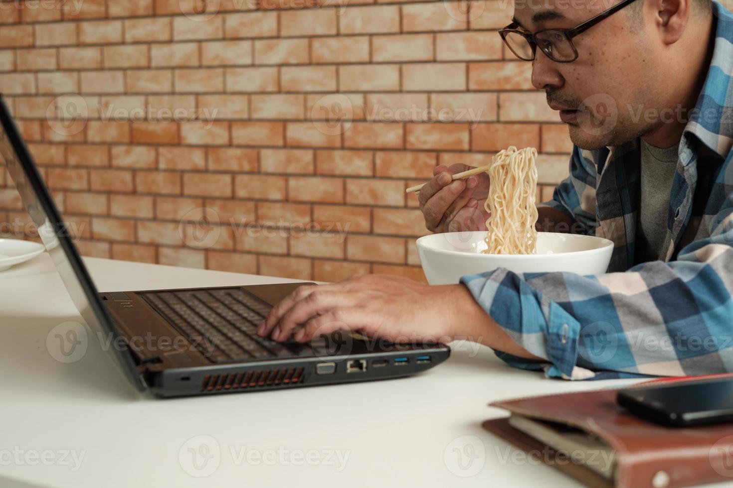 trabalhador tailandês do sexo masculino ocupado trabalhando com laptop, use os pauzinhos para comer macarrão instantâneo apressadamente durante o intervalo do almoço do escritório, porque é rápido, saboroso e barato. ao longo do tempo fast food asiático, estilo de vida pouco saudável. foto