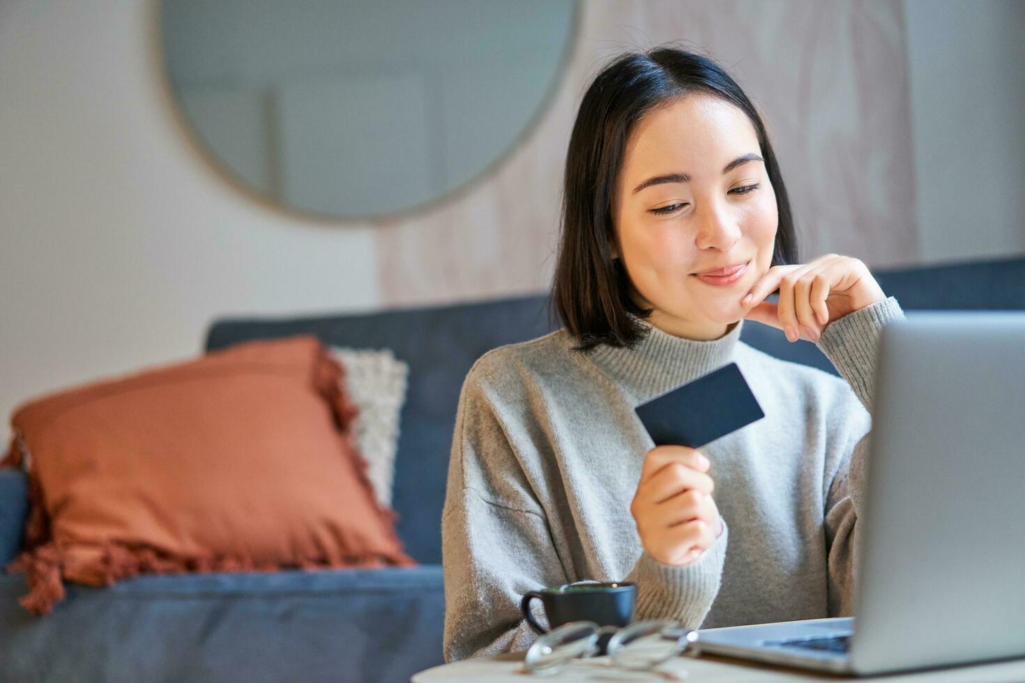 feliz sorridente menina com crédito cartão, pagando dela contas conectados em computador, fazendo compras em dela computador portátil, sentado às casa foto