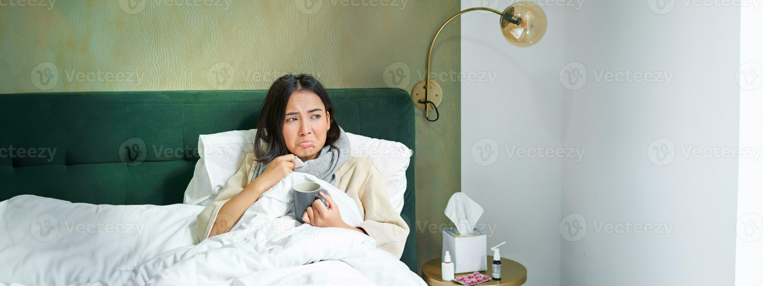 COVID-19, gripe e vacinação conceito. coreano menina deitado dentro cama com frio, pegando gripe, bebendo quente chá e usando prescrito medicação foto