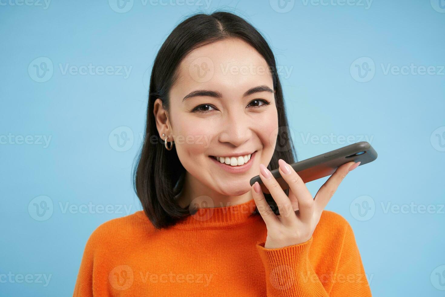 retrato do ásia mulher registros voz mensagem, fala em viva voz, detém Móvel telefone perto boca enquanto Falando, traduz dela discurso em aplicativo, azul fundo foto