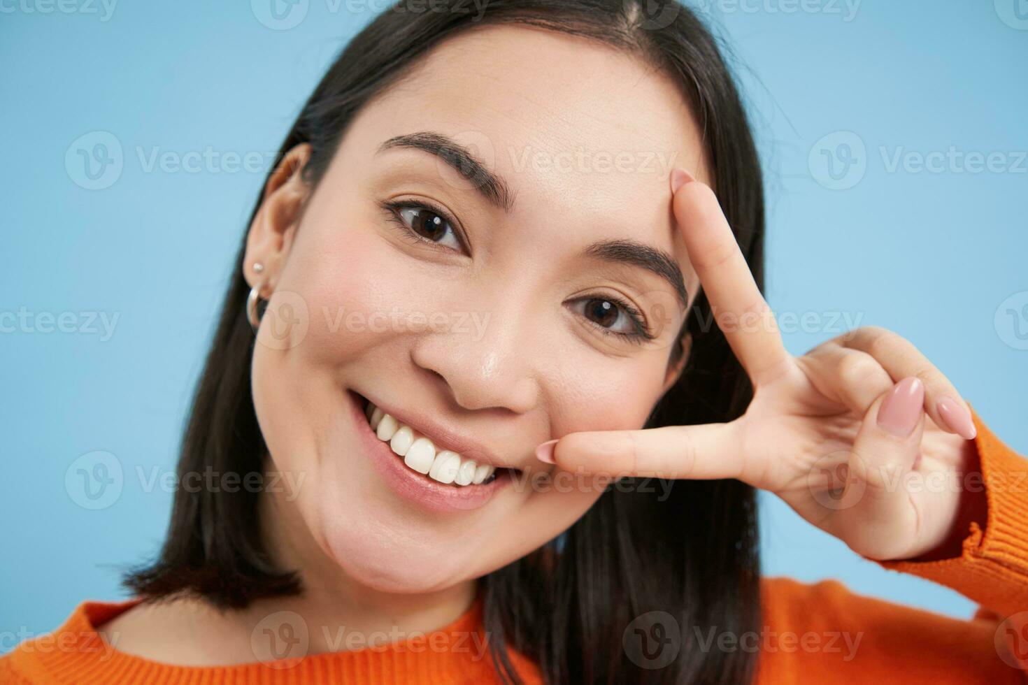 positivo e feliz japonês mulher, mostra Paz placa e sorrisos, feliz sinal de v gesto, em pé sobre azul fundo foto