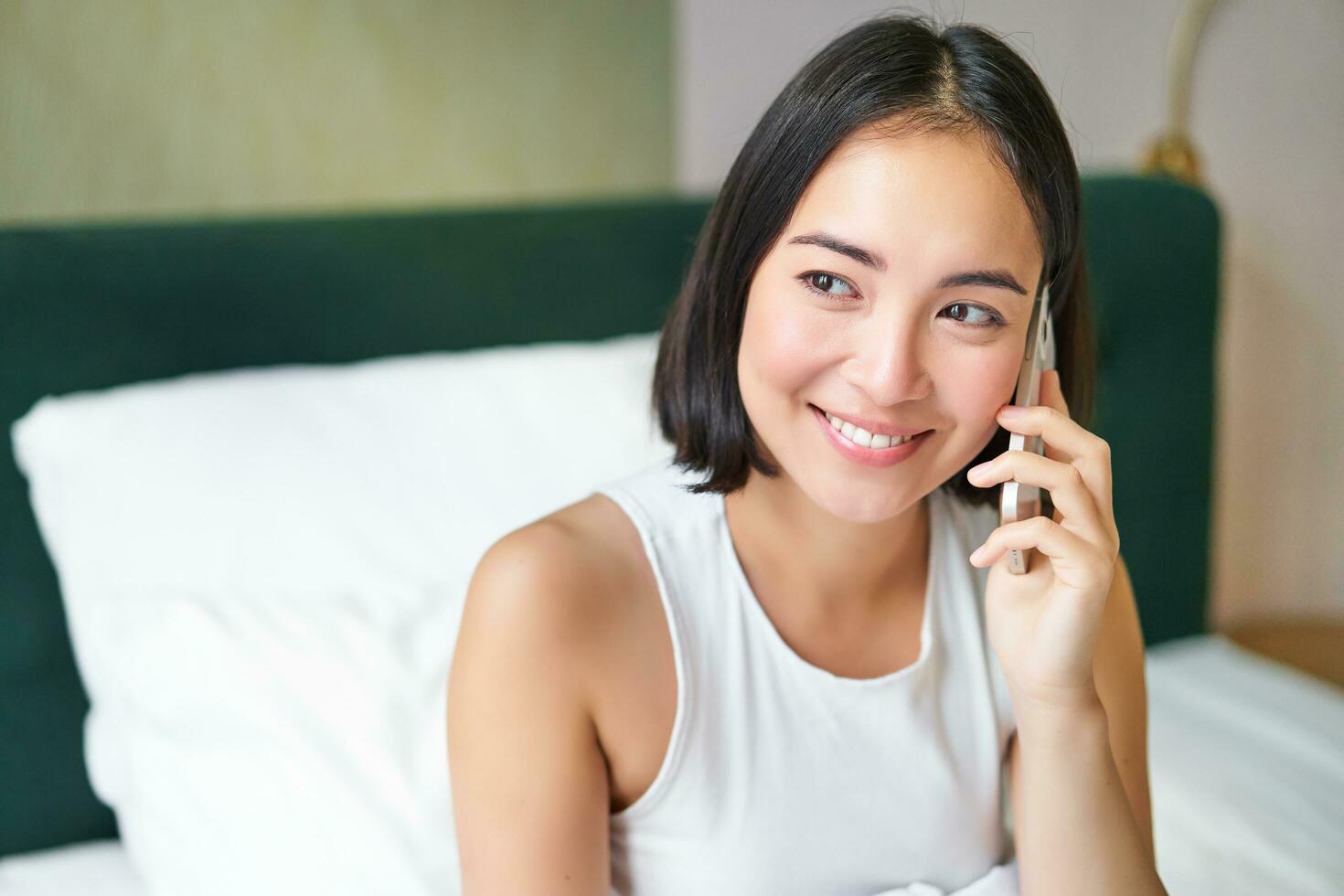 sorridente coreano menina dentro cama, fala em Móvel telefone, fazer uma telefone chamar, preguiçoso manhã Como ásia mulher ordens Entrega através da Smartphone foto