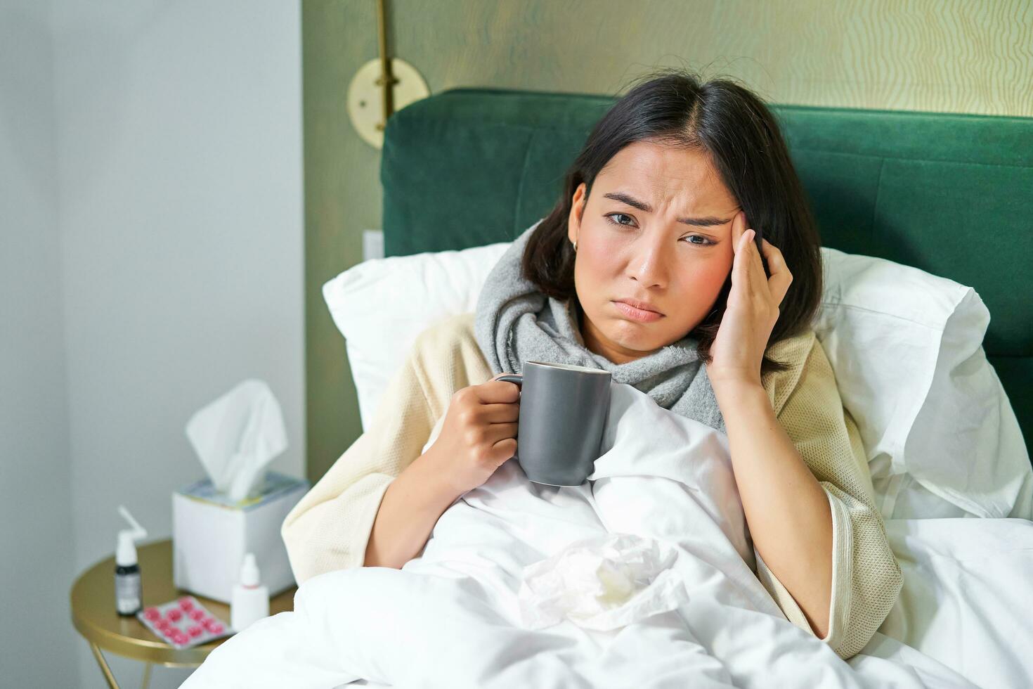 retrato do ásia mulher com dor de cabeça, pegando col, ficando em doente sair às lar, deitado dentro cama, bebendo quente chá, tendo gripe foto