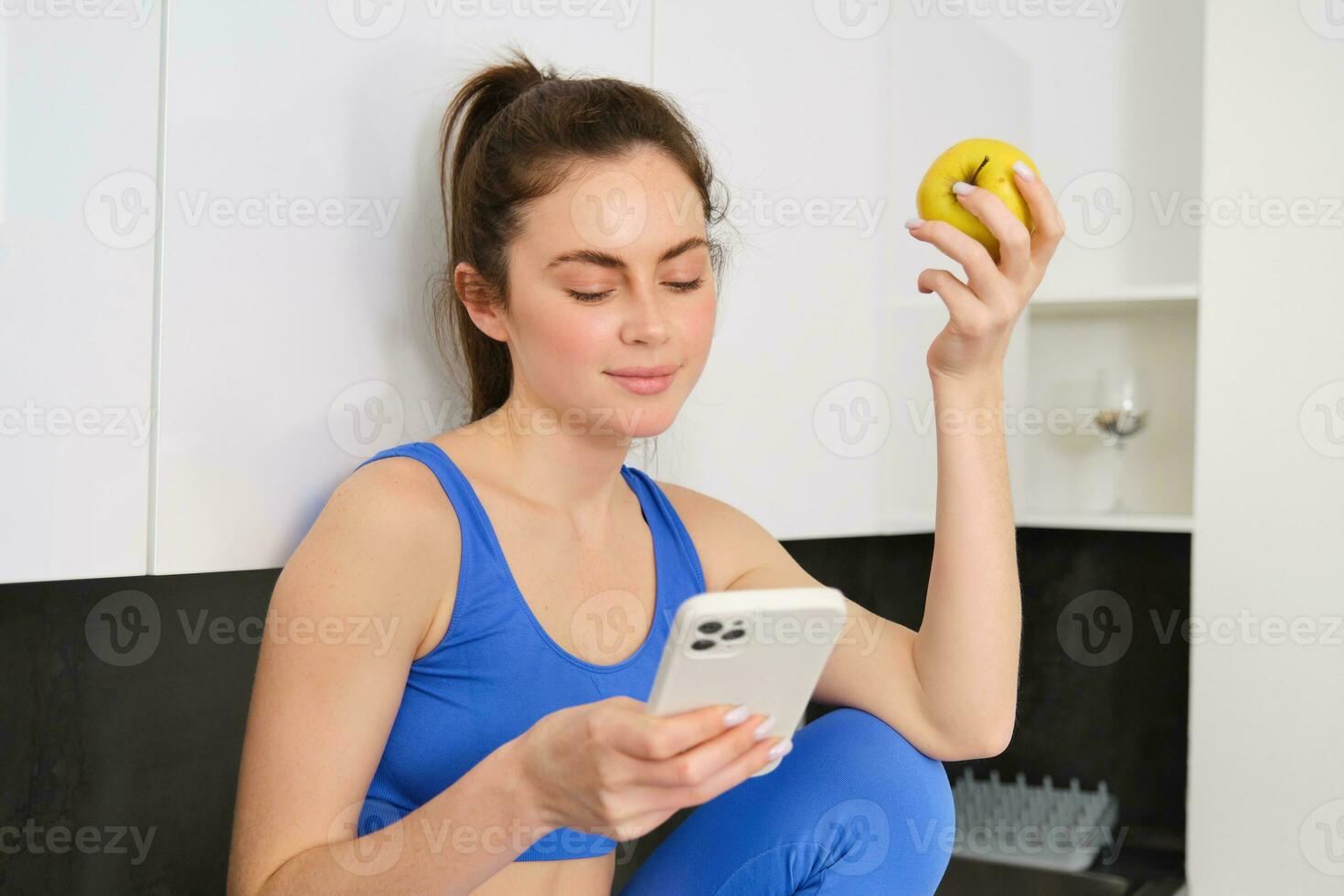 retrato do morena ginástica mulher, comendo a maçã, segurando Smartphone, usando Móvel telefone aplicativo enquanto tendo saudável fruta lanche dentro cozinha foto