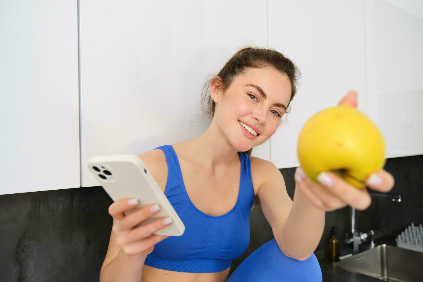 retrato do lindo, sorridente jovem ginástica mulher, oferta você a maçã, comendo saudável lanche, segurando uma fruta, sentado dentro cozinha foto