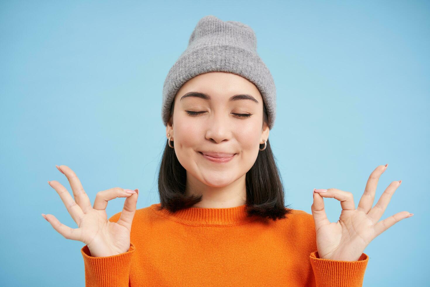 sons mente dentro saudável corpo. sorridente calma e relaxado ásia menina dentro gorro, mostra zen, relaxamento gesto, meditando, em pé sobre azul fundo foto