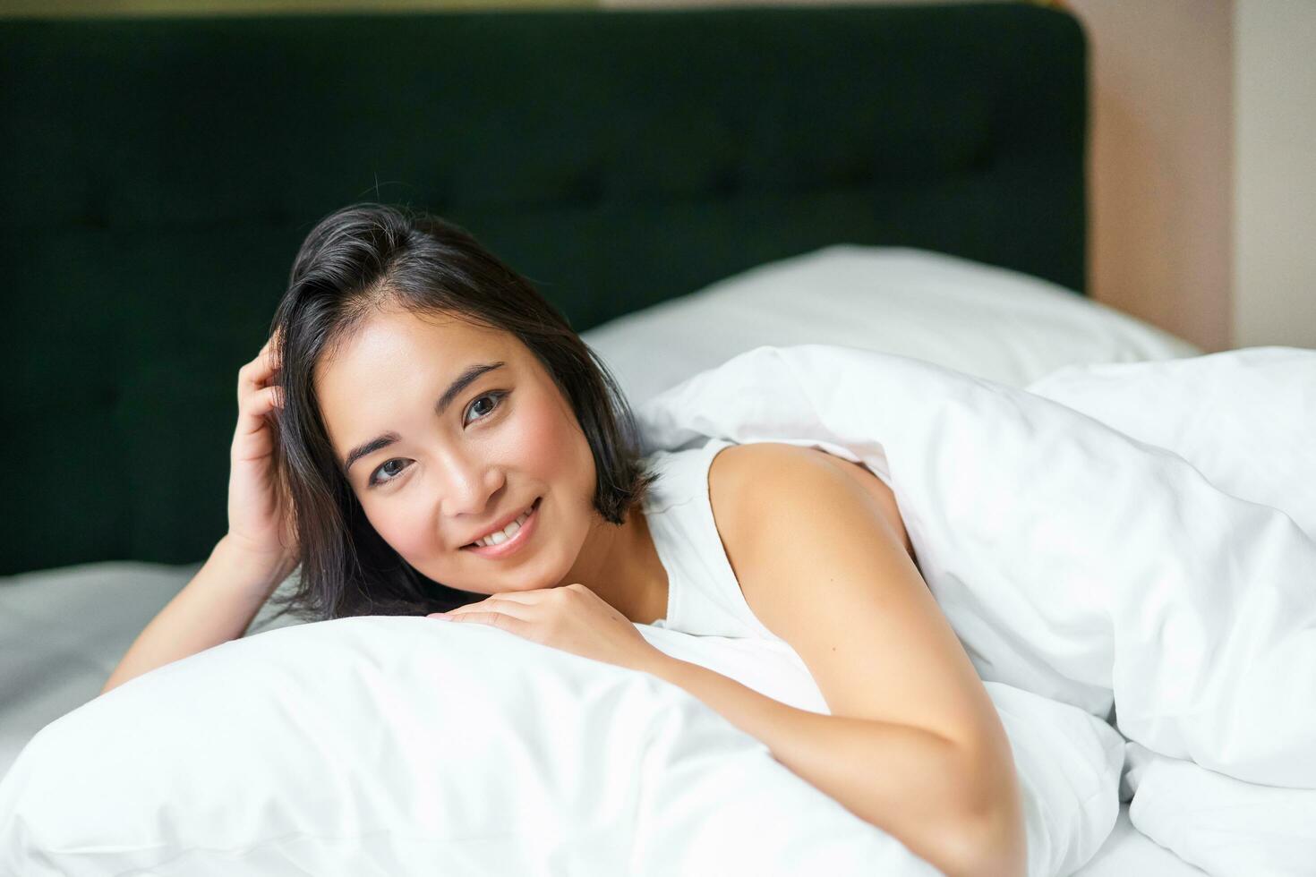 retrato do jovem coreano mulher sorridente, deitado dentro cama em travesseiro, posando dentro dela quarto, olhando relaxado e sentindo-me confortável foto