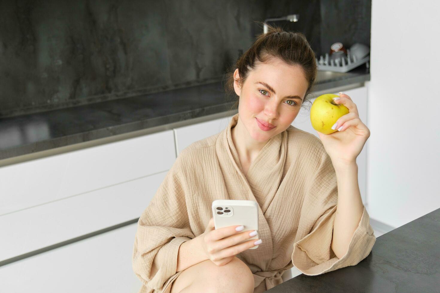 imagem do atraente jovem mulher dentro roupão de banho, senta dentro cozinha, come verde maçã e parece às Móvel telefone, usa Smartphone aplicativo, ordens Comida em inscrição foto