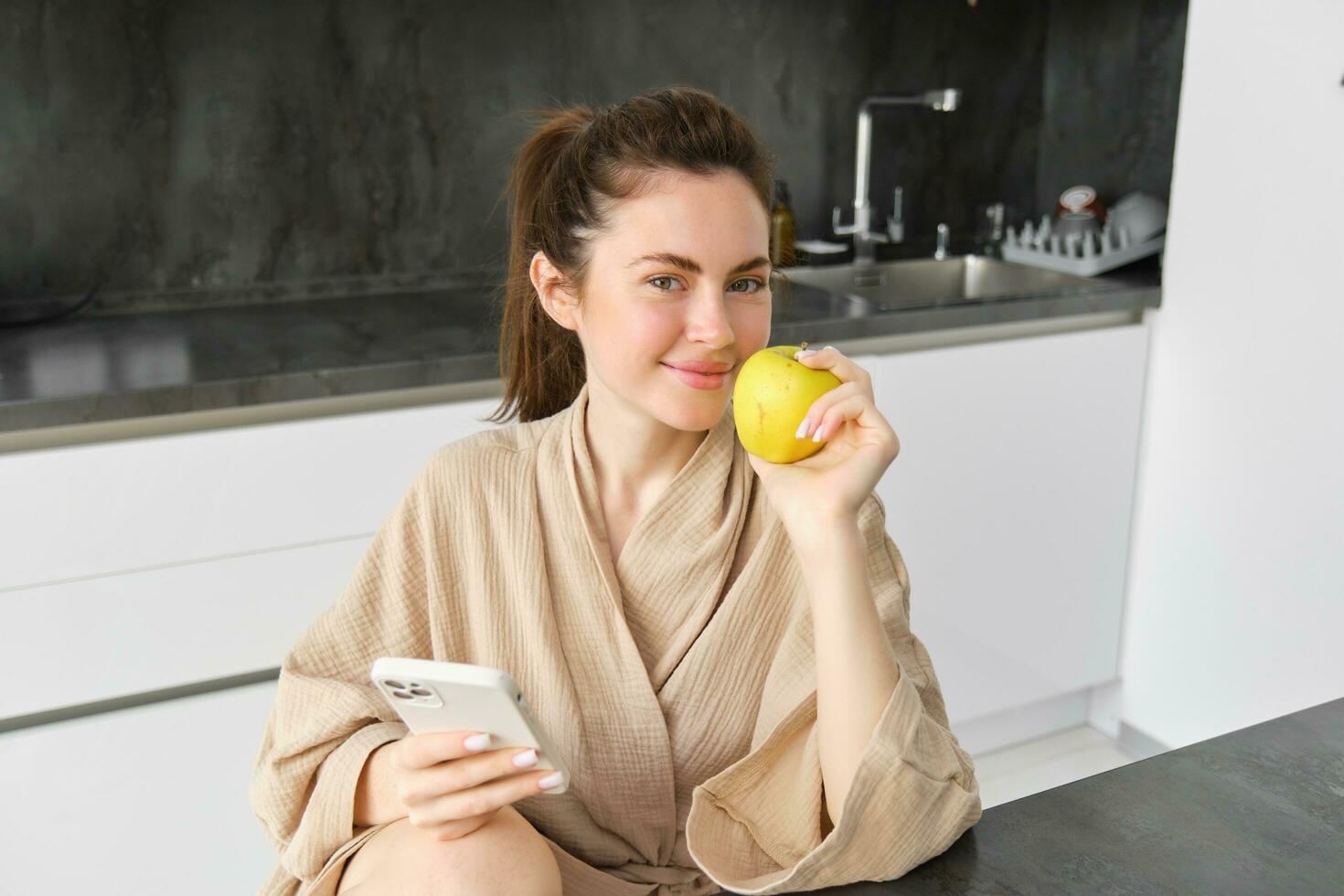 imagem do atraente jovem mulher dentro roupão de banho, senta dentro cozinha, come verde maçã e parece às Móvel telefone, usa Smartphone aplicativo, ordens Comida em inscrição foto