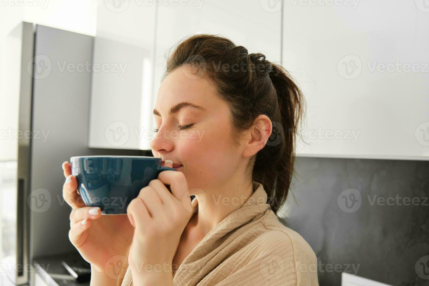 diariamente rotina e estilo de vida. jovem lindo mulher dentro roupão de banho, em pé dentro cozinha com copo do café, bebendo chá, sorridente e olhando feliz foto