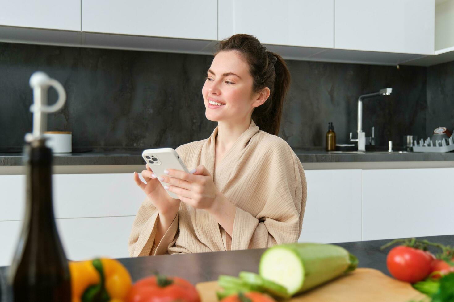 retrato do lindo jovem mulher com Smartphone, sentado dentro a cozinha, cozinhando jantar, legumes em cortar borda. menina assistindo cozinhando receita em Móvel telefone foto