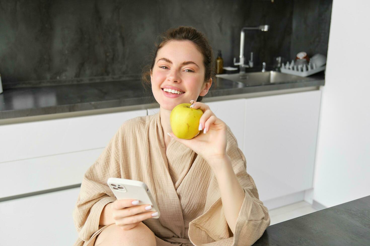 fechar acima retrato do feliz jovem mulher dentro roupão de banho, sentado dentro a cozinha e usando Móvel telefone, segurando a maçã, ordem frutas e legumes on-line, usando Smartphone aplicativo para mercearias Entrega foto