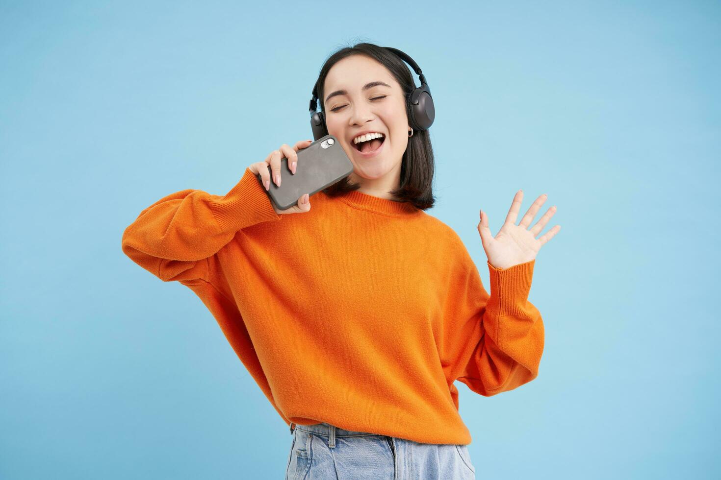 lindo coreano menina cantando dentro Smartphone microfone, escuta música dentro fones de ouvido, canta karaokê em Móvel aplicativo, carrinhos sobre azul fundo foto