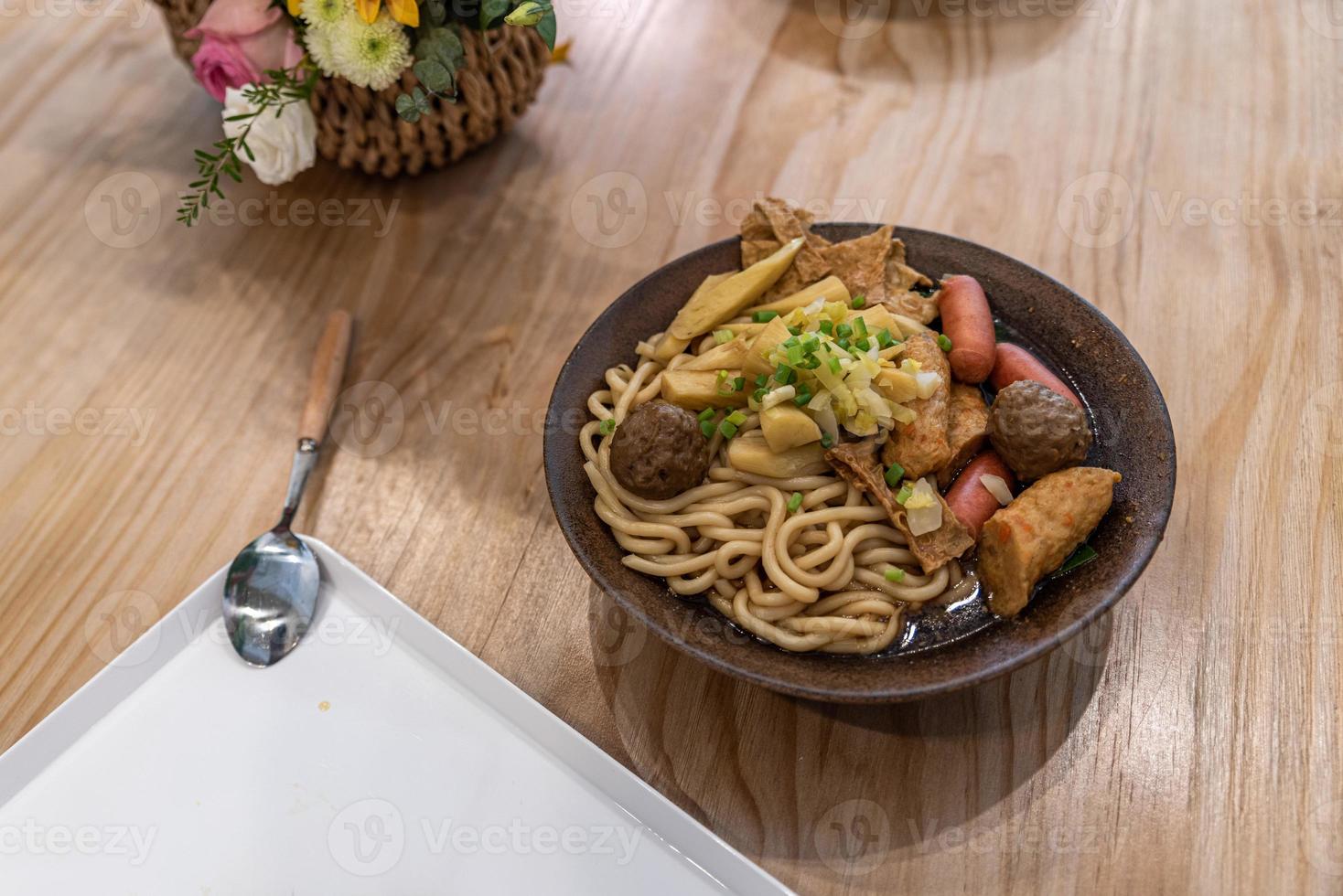 uma tigela de sopa de macarrão com salsicha, legumes e carne está na mesa foto