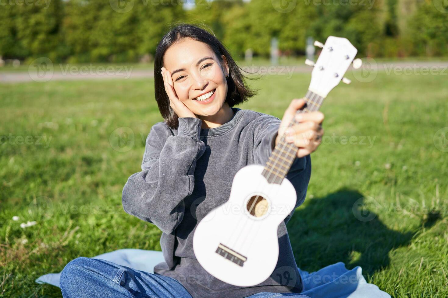 lindo ásia menina com feliz sorriso, mostra dela cavaquinho, senta lado de fora dentro parque em grama, relaxa com música foto