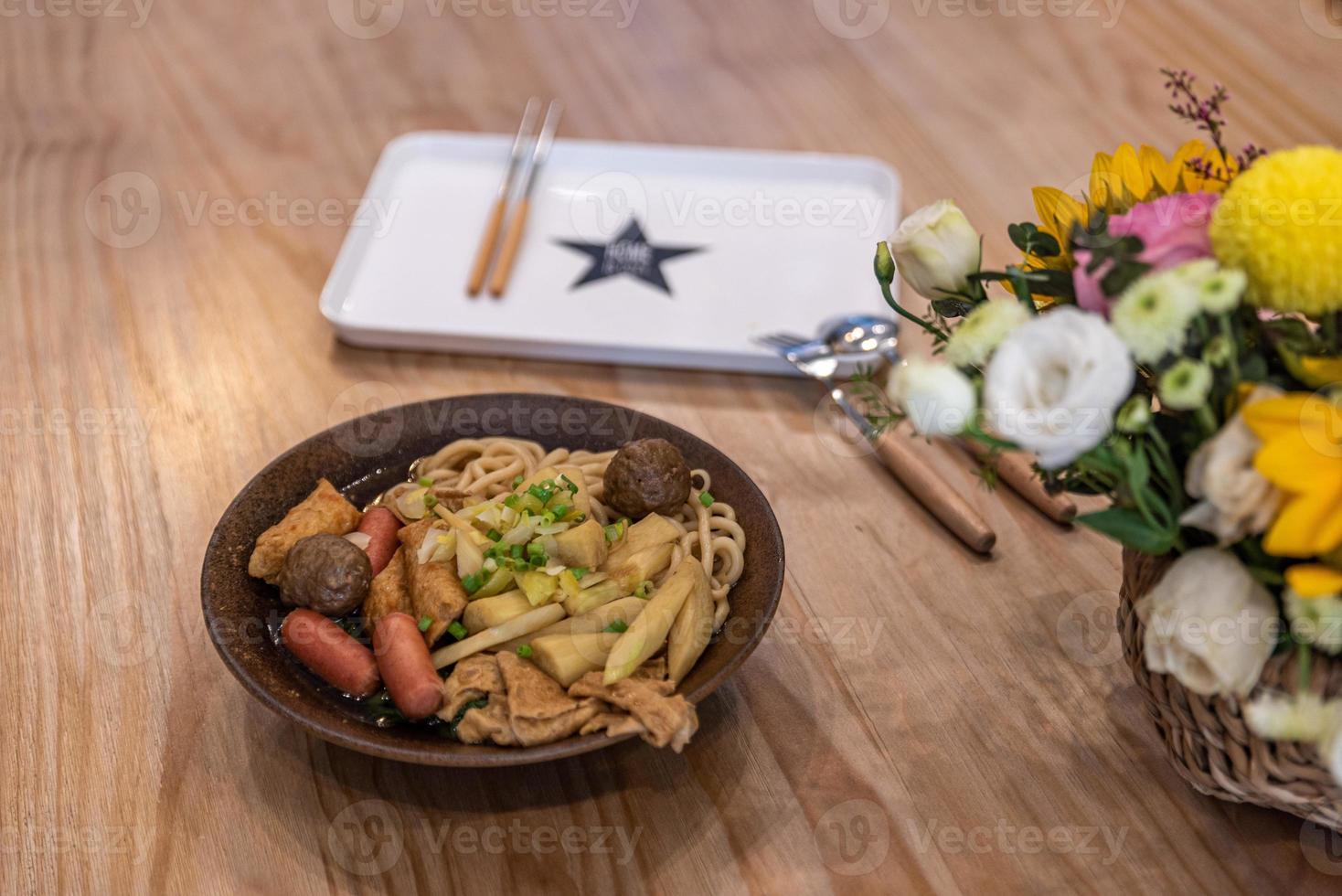 uma tigela de sopa de macarrão com salsicha, legumes e carne está na mesa foto