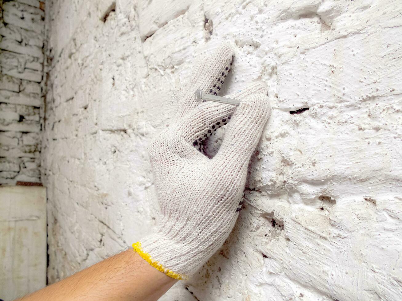 uma homem é segurando uma unha para estar bater em uma branco tijolo parede usando branco luvas foto