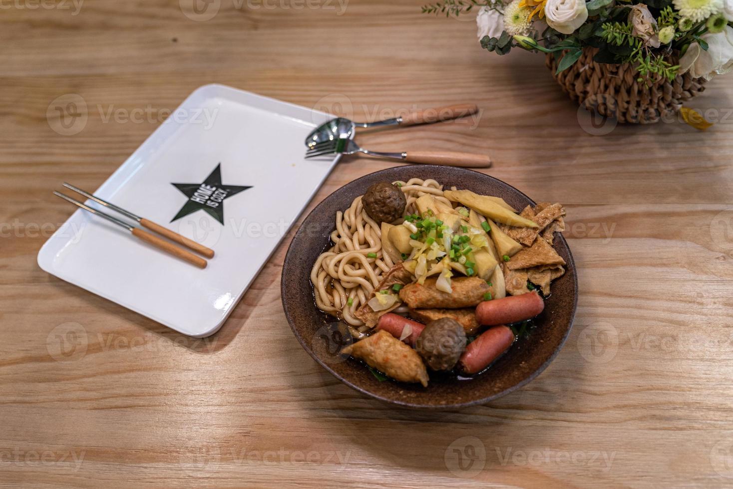 uma tigela de sopa de macarrão com salsicha, legumes e carne está na mesa foto