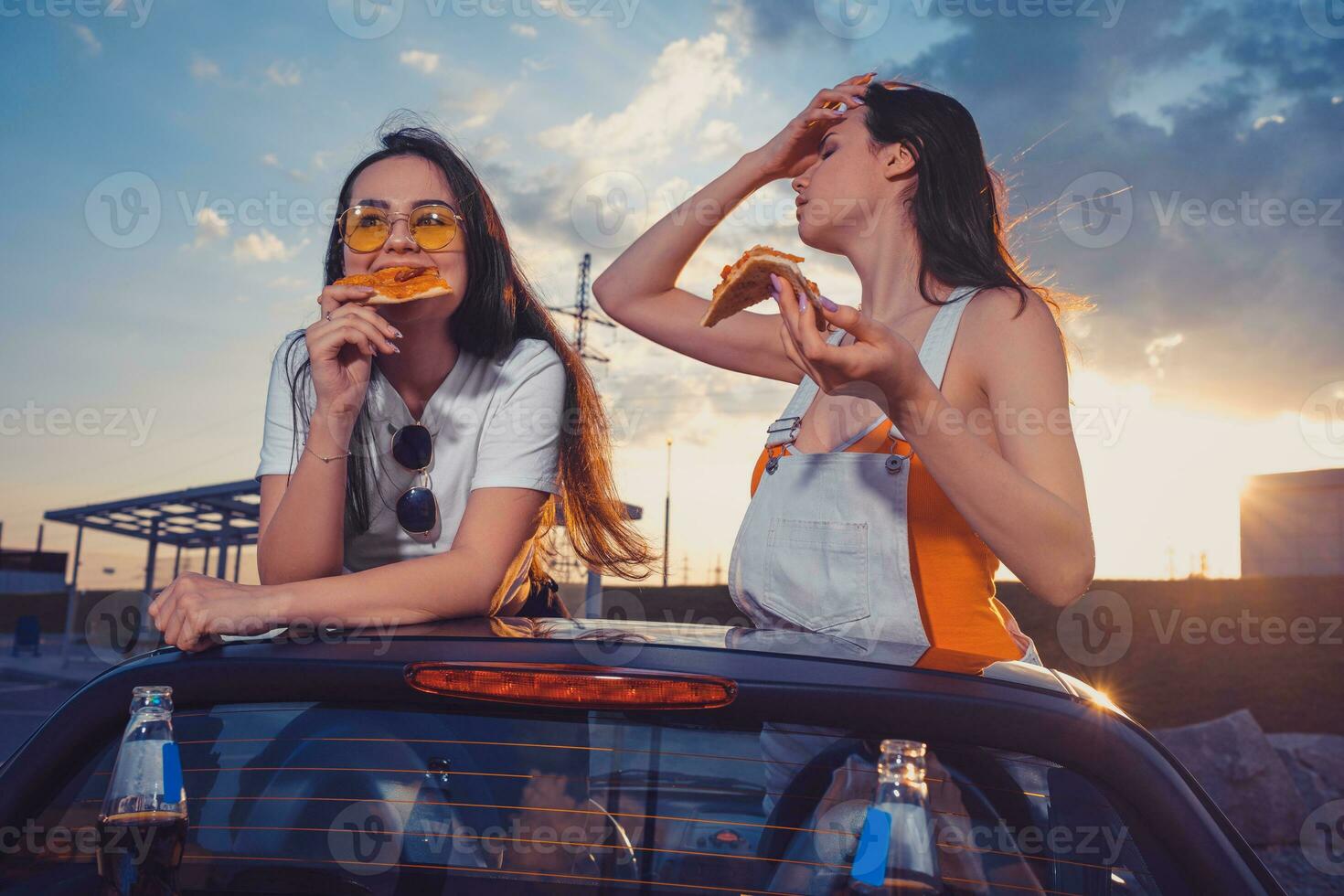 feliz modelos dentro casual equipamento estão comendo pizza, posando dentro amarelo carro cavalo estradeiro com refrigerante água dentro vidro garrafas em Está porta-malas. velozes Comida. cópia de espaço foto