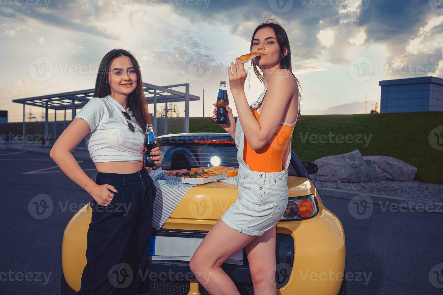 amigos fêmeas dentro casual equipamento estão sorridente, comendo pizza, desfrutando refrigerante dentro vidro garrafas, posando perto amarelo carro em estacionamento. fechar acima, cópia de espaço foto