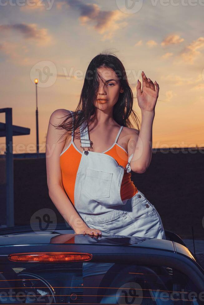 sexy jovem fêmea dentro branco No geral e laranja camiseta é posando ao ar livre em pé dentro carro conversível. verão noite, pôr do sol. fechar acima foto