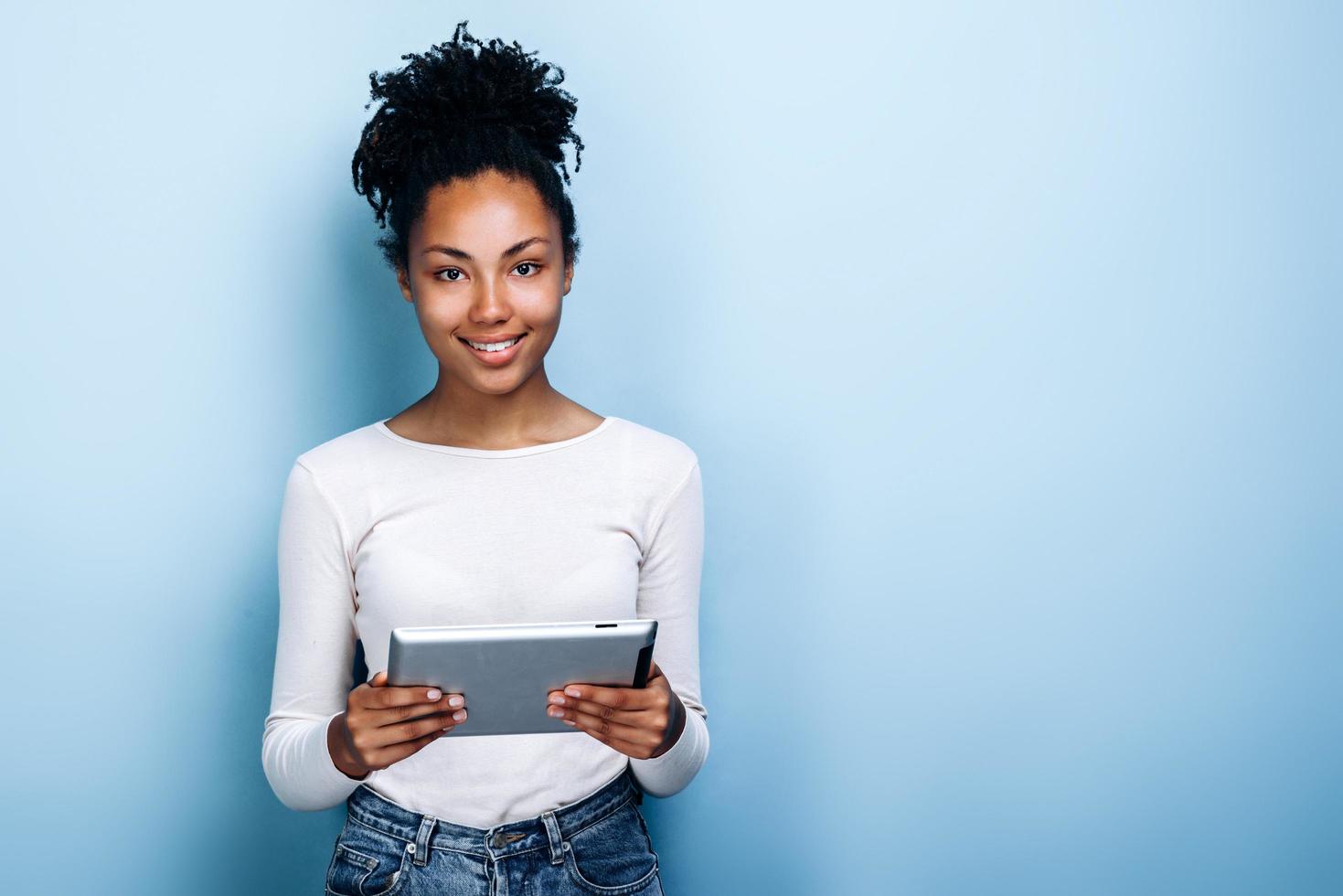 Mulher jovem e bonita com cabelo encaracolado está com um tablet nas mãos sobre um fundo de parede azul foto