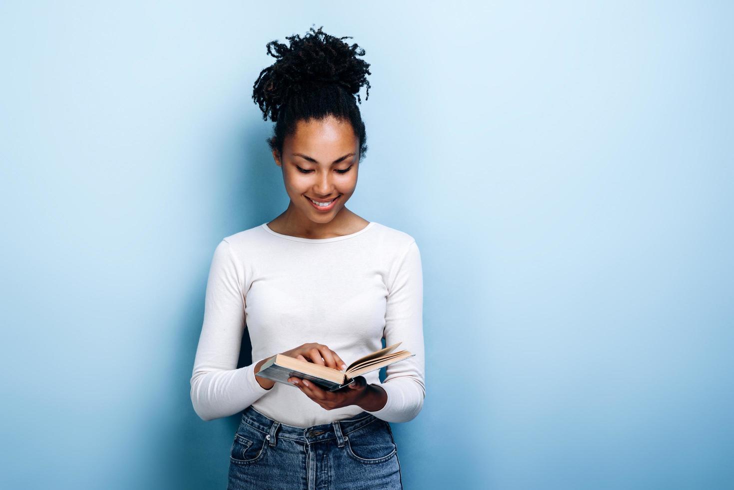 Mulher jovem e atraente com um sorriso lendo um livro interessante sobre um fundo de parede azul foto